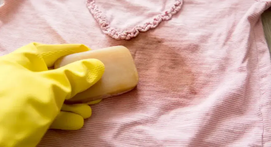 Gelbe Handschuhe halten Gallseife, um einen Fleck auf einem rosafarbenen gestreiften Shirt zu behandeln.
