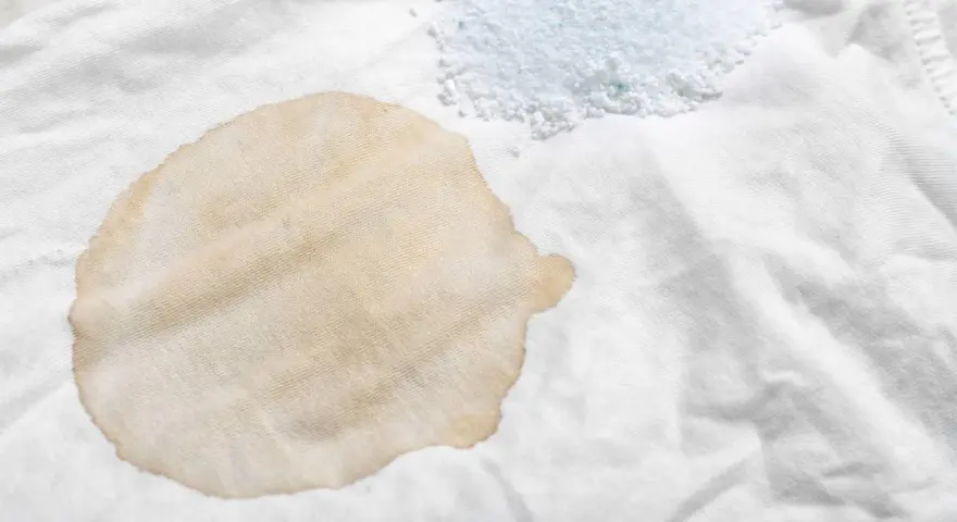 Ein brauner Fleck auf weißem Stoff, dane ein Haufen Waschpulver. So lassen sich hartnäckige teeflecken entfernen.