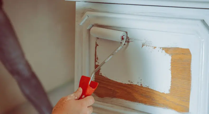 Eine Hand mit einem Farbroller trägt weiße Farbe auf ein Holzmöbel auf. Das Holz wird vorbereitet, um es zu entfetten und neu zu streichen.