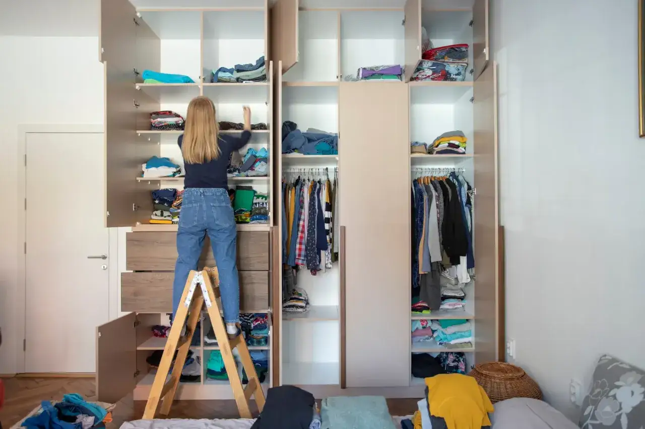 M&auml;dchen auf Trittleiter sortiert Kleidung im offenen Kleiderschrank, um Ordnung zu halten.