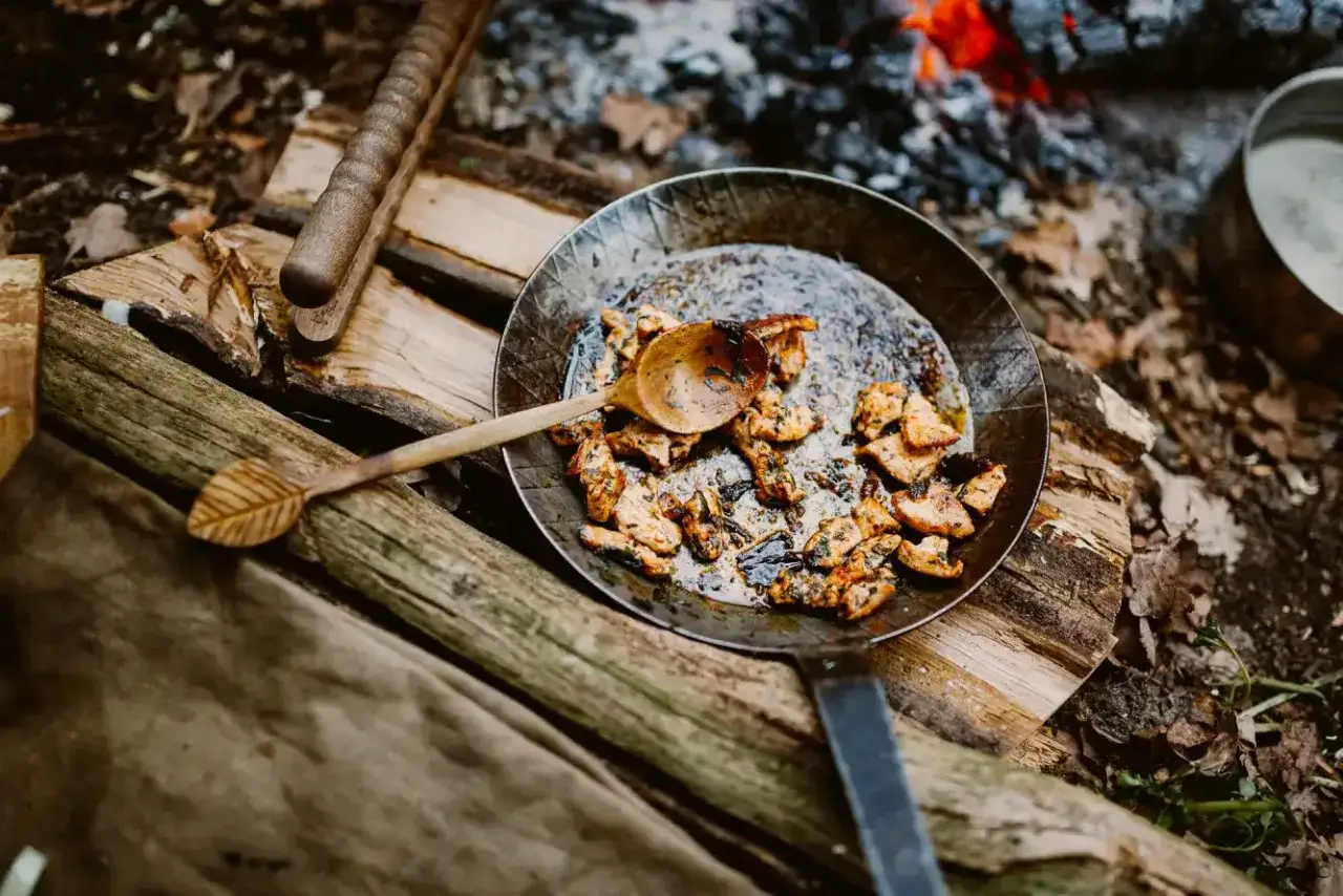 Gusseisenpfanne reinigen: Gebratenes Fleisch in der Pfanne &uuml;ber offenem Feuer, daneben ein Holzl&ouml;ffel.