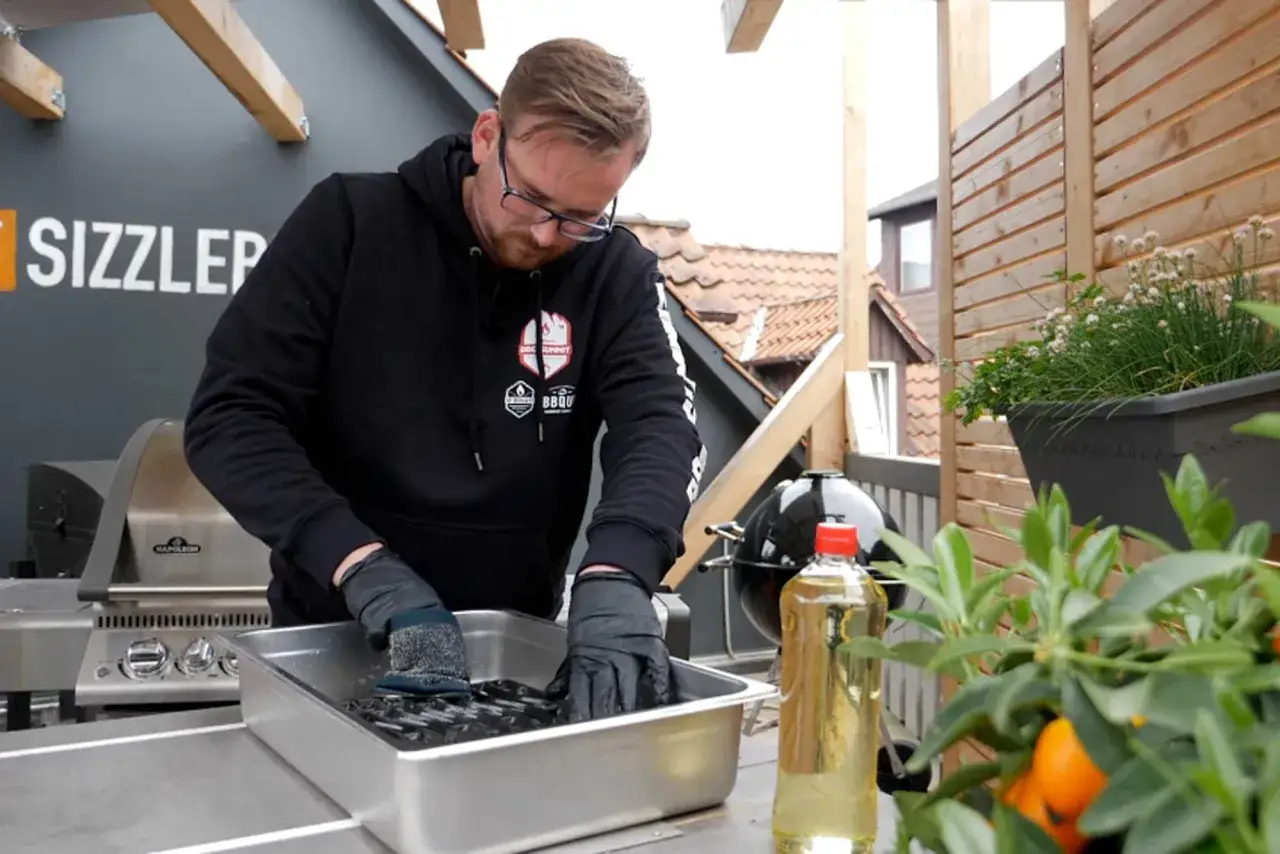 Mann reinigt emaillierte Grillroste in einer Wanne. Neben ihm steht eine Flasche &Ouml;l und ein Grill.