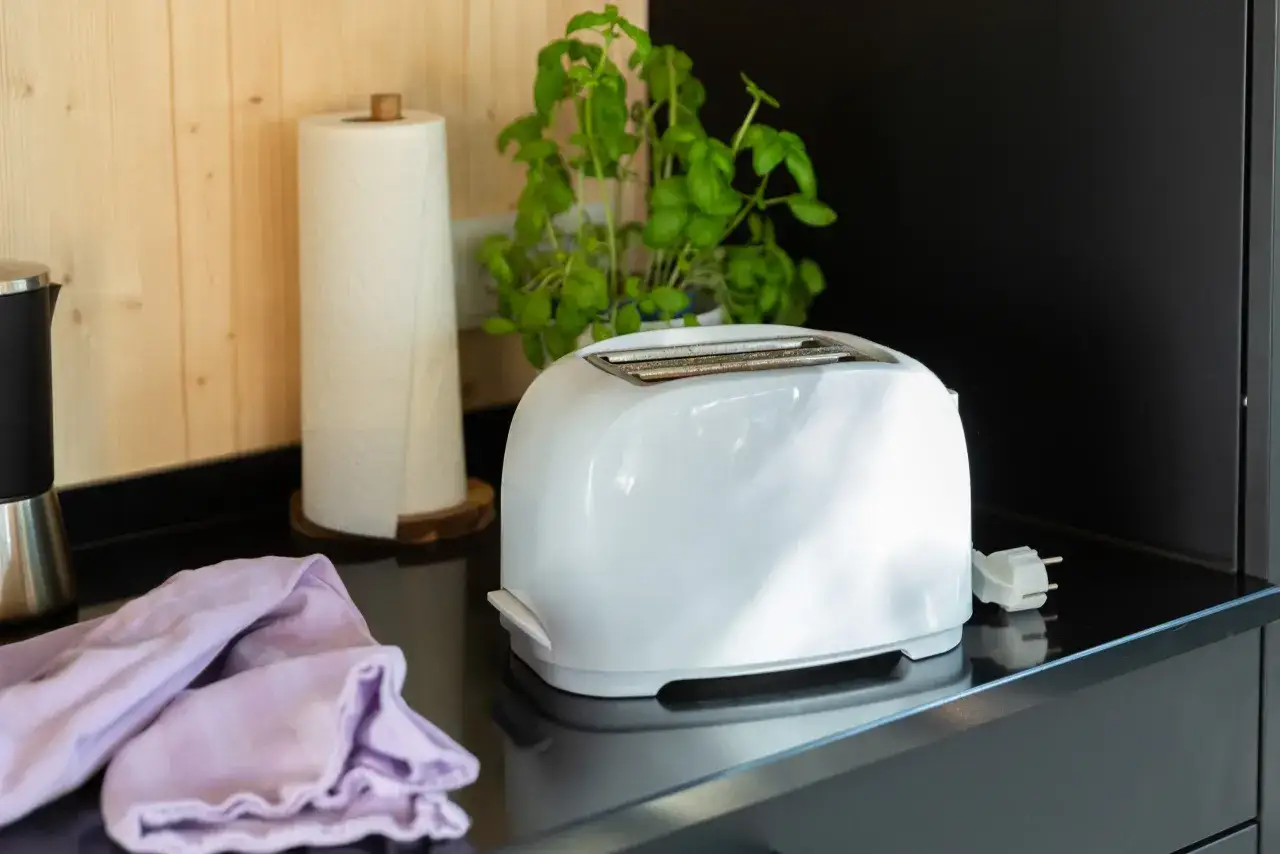 Ein wei&szlig;er Toaster steht auf einer schwarzen Arbeitsplatte, bereit zum toaster reinigen. Daneben liegen ein K&uuml;chentuch und eine K&uuml;chenrolle.