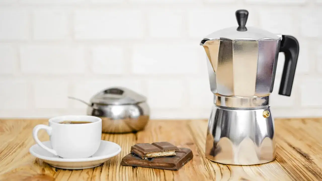 Ein gl&auml;nzender Espressokocher steht neben einer Tasse Kaffee und Schokolade. Nach dem Genuss ist das espressokocher reinigen wichtig.