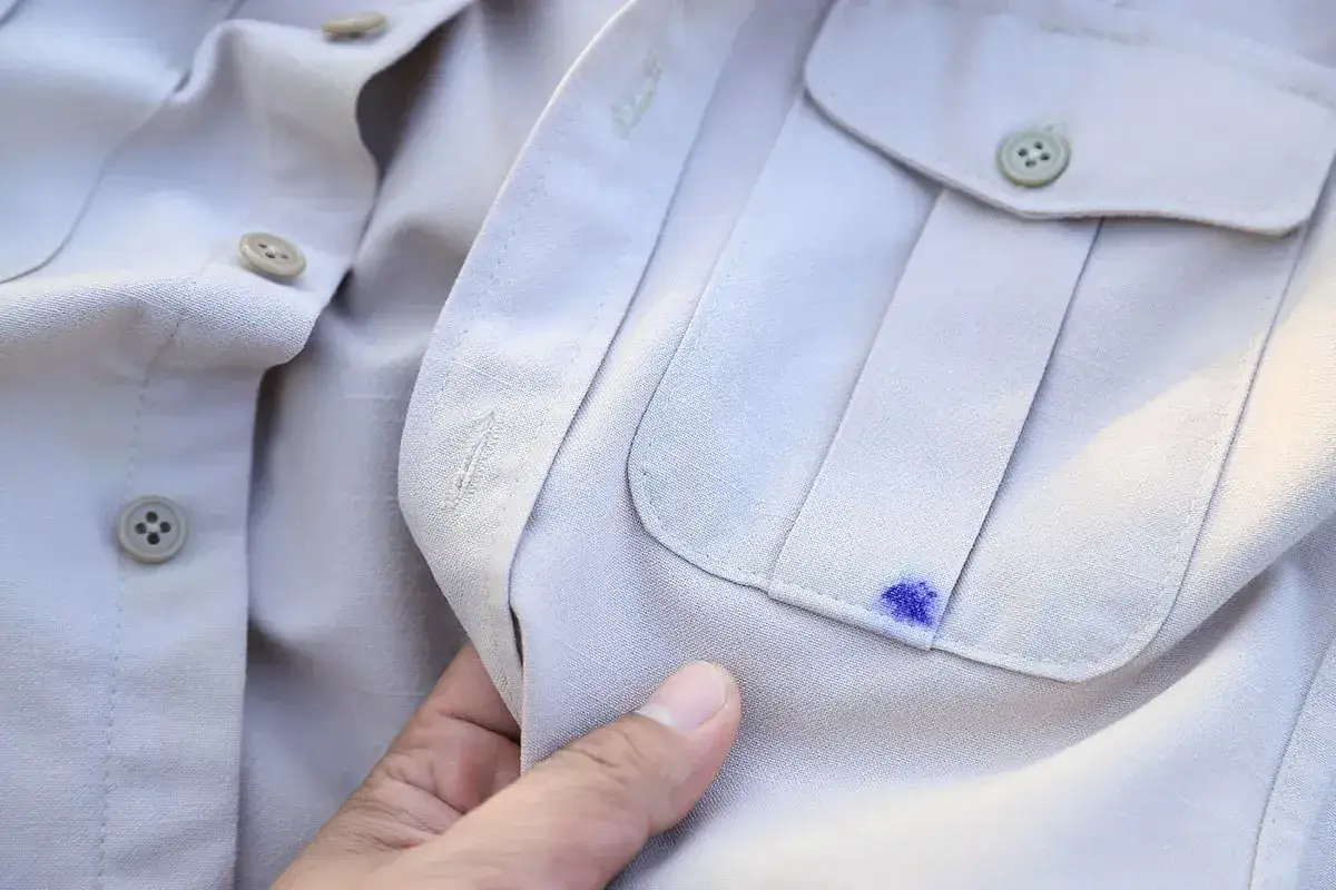 Ein blauer Tintenfleck auf einem hellen Hemd. Ein Finger berührt den Fleck, um zu zeigen, wie man Tinte aus Kleidung entfernen kann.