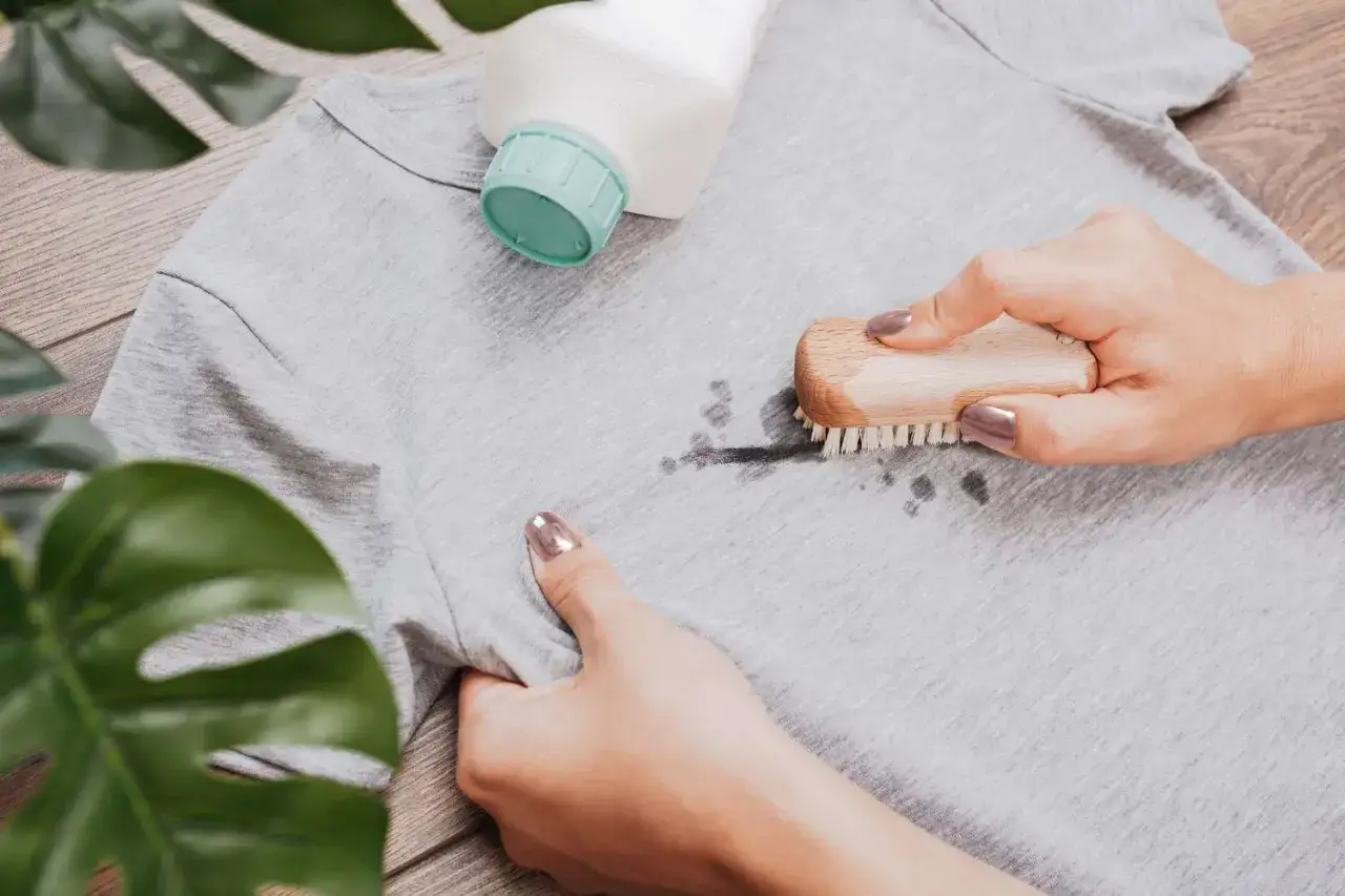 Mascara aus Kleidung entfernen: Mit einer B&uuml;rste und Reiniger wird ein Fleck auf einem grauen T-Shirt bearbeitet.
