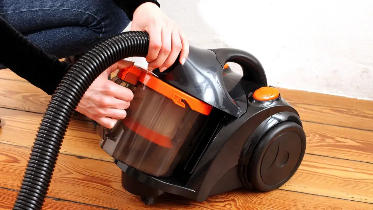 H&auml;nde reinigen den Staubbeh&auml;lter eines schwarzen Staubsaugers mit orangefarbenen Akzenten auf Holzboden.