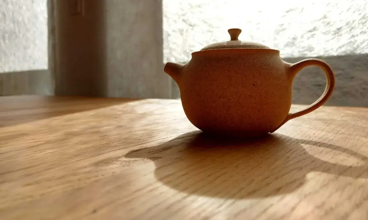 Eine kleine, erdbeerfarbene Teekanne steht auf einem Holztisch. Das Sonnenlicht f&auml;llt durch ein Fenster und wirft Schatten. Zeit, die Teekanne zu reinigen.