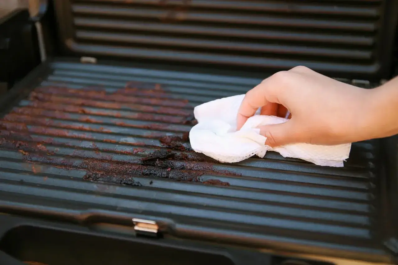 Hand wischt mit K&uuml;chenpapier &uuml;ber die verkrusteten Rillen eines Elektrogrills, um ihn zu reinigen.
