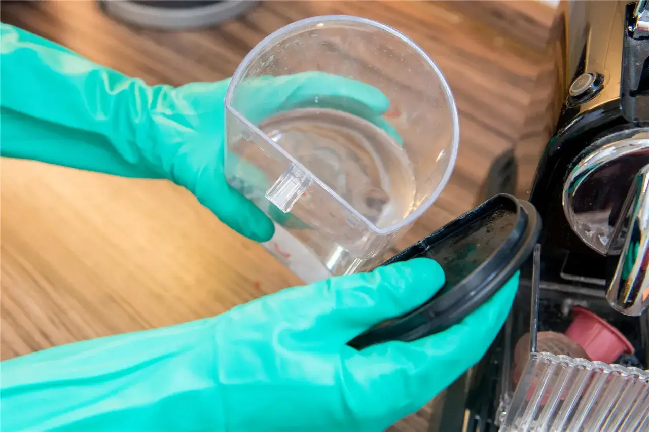 Mit gr&uuml;nen Handschuhen wird eine Kaffeemaschine entkalkt. Ein Beh&auml;lter mit Fl&uuml;ssigkeit wird &uuml;ber dem Auffangbeh&auml;lter gehalten.
