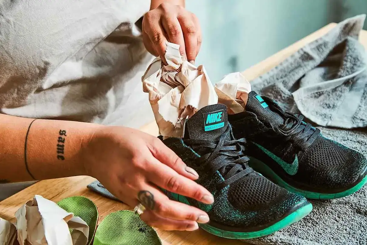 H&auml;nde stopfen Papier in schwarze Nike-Schuhe, um sie zu trocknen. Ein Handtuch liegt daneben.