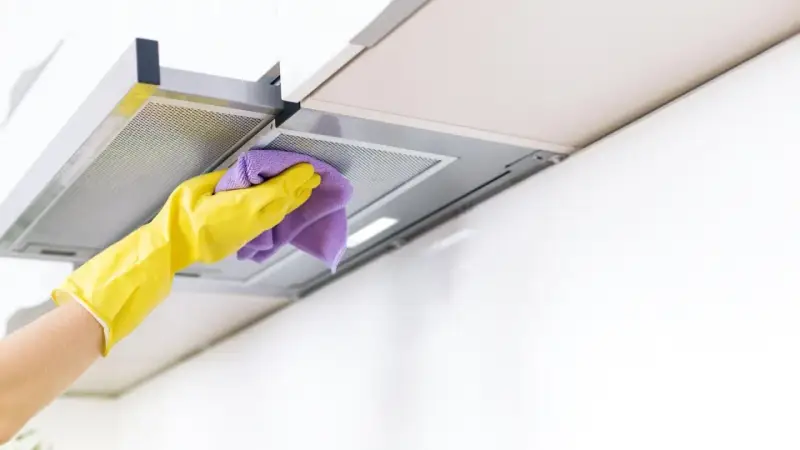 Gelbe Handschuhe wischen mit einem lila Tuch über die Dunstabzugshaube. Alu reinigen für strahlenden Glanz in der Küche.